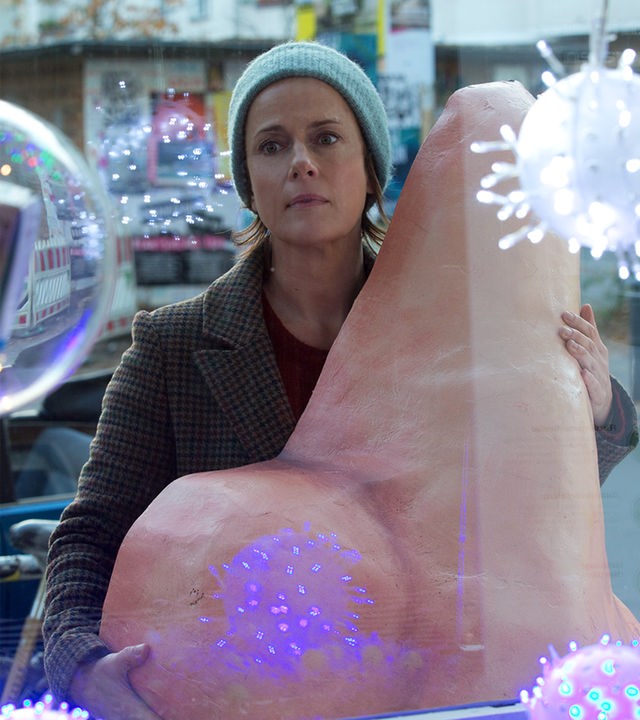 Vor dem Schaufenster einer Apotheke steht Tanja (Claudia Michelsen) mit einer Pappmaché-Nase. Im Fenster blinken bunte Lichtkugeln, die wie große Viren aussehen.