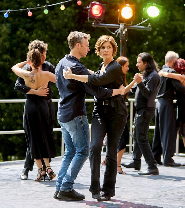 "In Wahrheit - Die Liebe und der Tod": Judith Mohn (Christina Hecke) und Freddy Breyer (Robin Sondermann) tanzen gemeinsam im Freien und werfen dabei suchende Blicke zur Seite. Hinter ihnen tanzen weitere Personen, umgeben von Grün und einer bunten Lichterkette.