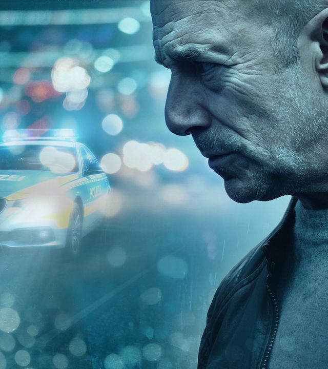Die Hauptfigur Ingo Thiel hat den Blick ernst nach unten gerichtet. Im Hintergrund steht ein Polizeiauto.