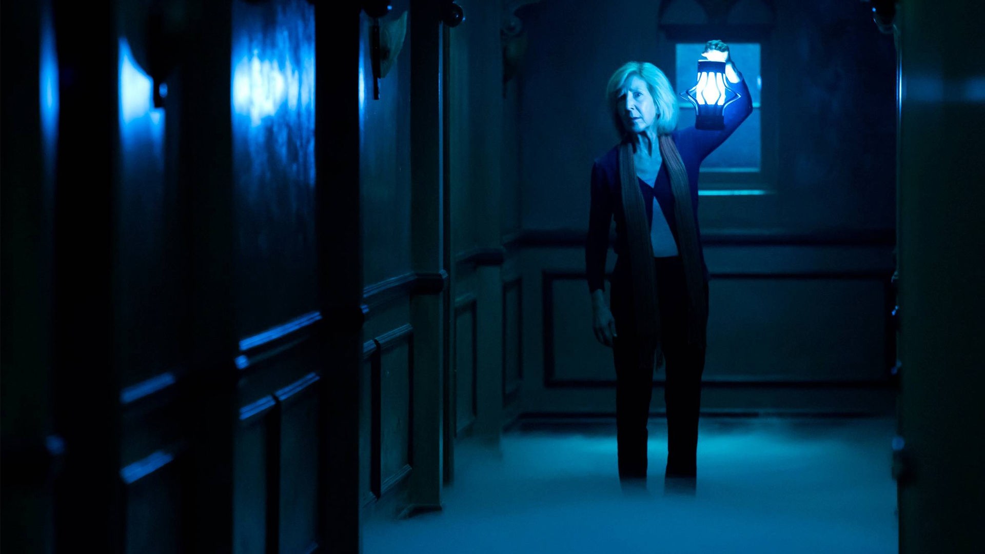 "Insidious: Chapter 3 - Jede Geschichte hat einen Anfang": Elise Rainier (Lin Shaye) steht in einem düsteren, nebligen Flur und blickt besorgt nach vorne. Sie trägt dunkle Kleidung und hält eine hell leuchtende, altmodische Laterne in die Höhe, die die Szene in kaltes, blaues Licht taucht.