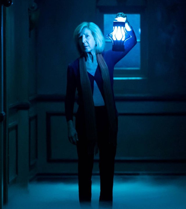 "Insidious: Chapter 3 - Jede Geschichte hat einen Anfang": Elise Rainier (Lin Shaye) steht in einem düsteren, nebligen Flur und blickt besorgt nach vorne. Sie trägt dunkle Kleidung und hält eine hell leuchtende, altmodische Laterne in die Höhe, die die Szene in kaltes, blaues Licht taucht.