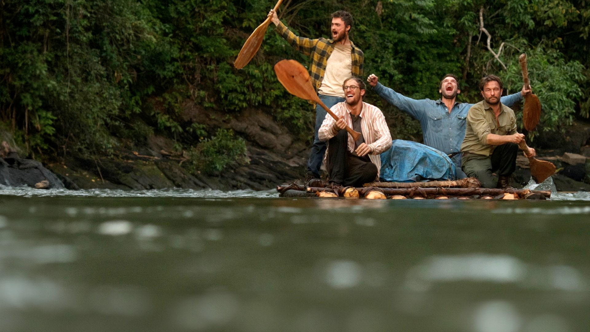 Yossi (Daniel Radcliffe), Marcus (Joel Jackson), Kevin (Alex Russell), alle mit ausgestreckten Armen jubelnd, und Karl (Thomas Kretschmann) sitzen auf einem, im Fluss treibenden, Floß.
