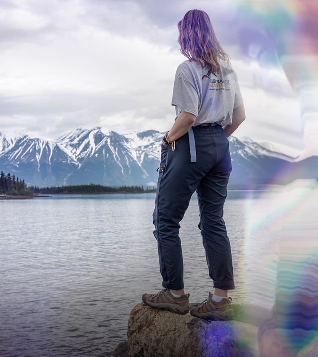 Eine Frau in der Rückenansicht steht auf einem Felsen und überblickt einen See in dessen Mitte sich eine Insel mit Nadelbäumen befindet.