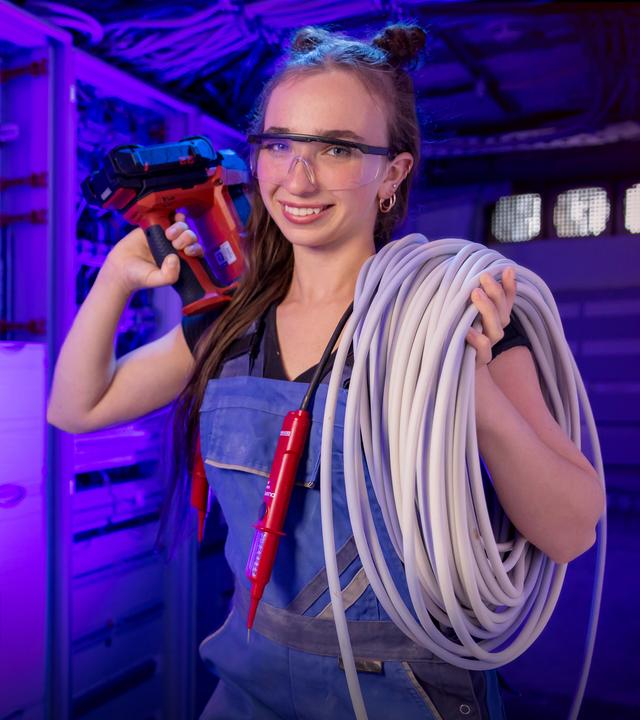 Eine Frau steht in blauer Arbeitskleidung in einem Technikraum. Sie hat eine Sicherheitsbrille an, hält eine Bohrmaschine in der einen Hand und ein aufgerolltes Kabel über der Schulter.