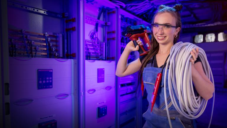 Eine Frau steht in blauer Arbeitskleidung in einem Technikraum. Sie hat eine Sicherheitsbrille an, hält eine Bohrmaschine in der einen Hand und ein aufgerolltes Kabel über der Schulter.