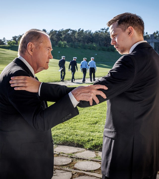 Martin (Peter Haber) und Vilhelm Beck (Valter Skarsgård) stehen sich im Freien gegenüber. Vilhelm legt seine linke Hand auf Martins Schulter, während Martin mit seiner rechten Hand Vilhelms Arm berührt. Im Hintergrund liegt eine große Grünanlage. Auf einem Gehweg laufen vier Polizisten (Komparsen) in Uniform.