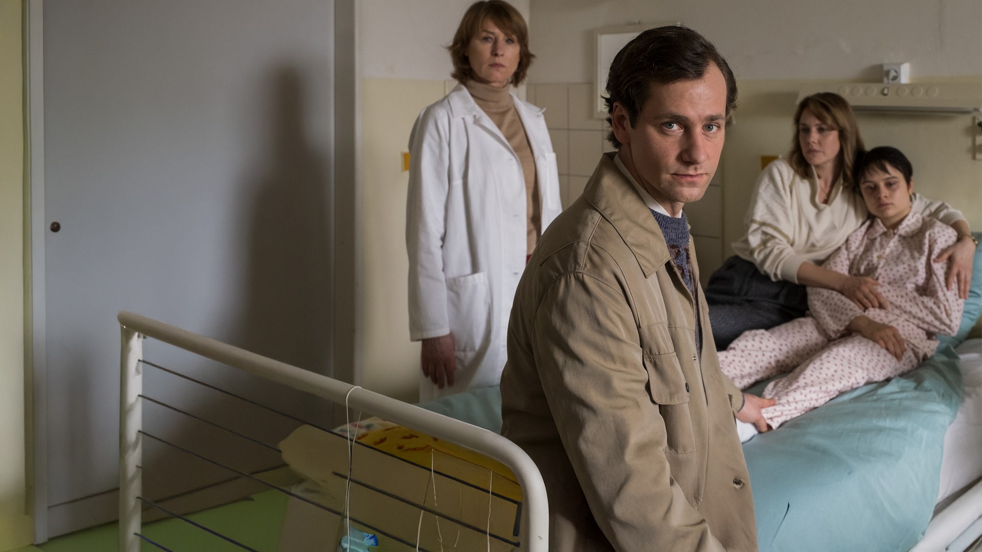  "Kranke Geschäfte": Armin Glaser (Florian Stetter) sitzt auf dem Krankenbett seiner Tochter Kati Glaser (Lena Urzendowsky) und schaut in die Kamera. Er greift mit einer Hand hinter sich an Katis Bein. Sie sitzt im Schlafanzug auf dem Bett, während ihre Mutter Marie Glaser (Felicitas Woll) sie fest im Arm hält. Neben dem Bett steht Dr. Sigurd (Corinna Harfouch), die mit starrem Blick in Armins Richtung schaut.