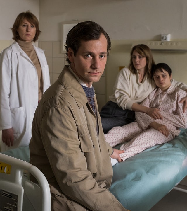  "Kranke Geschäfte": Armin Glaser (Florian Stetter) sitzt auf dem Krankenbett seiner Tochter Kati Glaser (Lena Urzendowsky) und schaut in die Kamera. Er greift mit einer Hand hinter sich an Katis Bein. Sie sitzt im Schlafanzug auf dem Bett, während ihre Mutter Marie Glaser (Felicitas Woll) sie fest im Arm hält. Neben dem Bett steht Dr. Sigurd (Corinna Harfouch), die mit starrem Blick in Armins Richtung schaut.