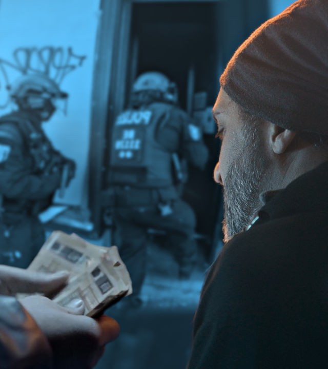 Im Hintergrund sind Polizisten in voller Ausrüstung zu sehen, die offenbar ein Haus stürmen. Im Vordergrund ist rechts ein Mann mit Bart und Mütze zu sehen. Links daneben sieht man die Hände eines anderen Mannes, der 50 Euro-Scheine hält.