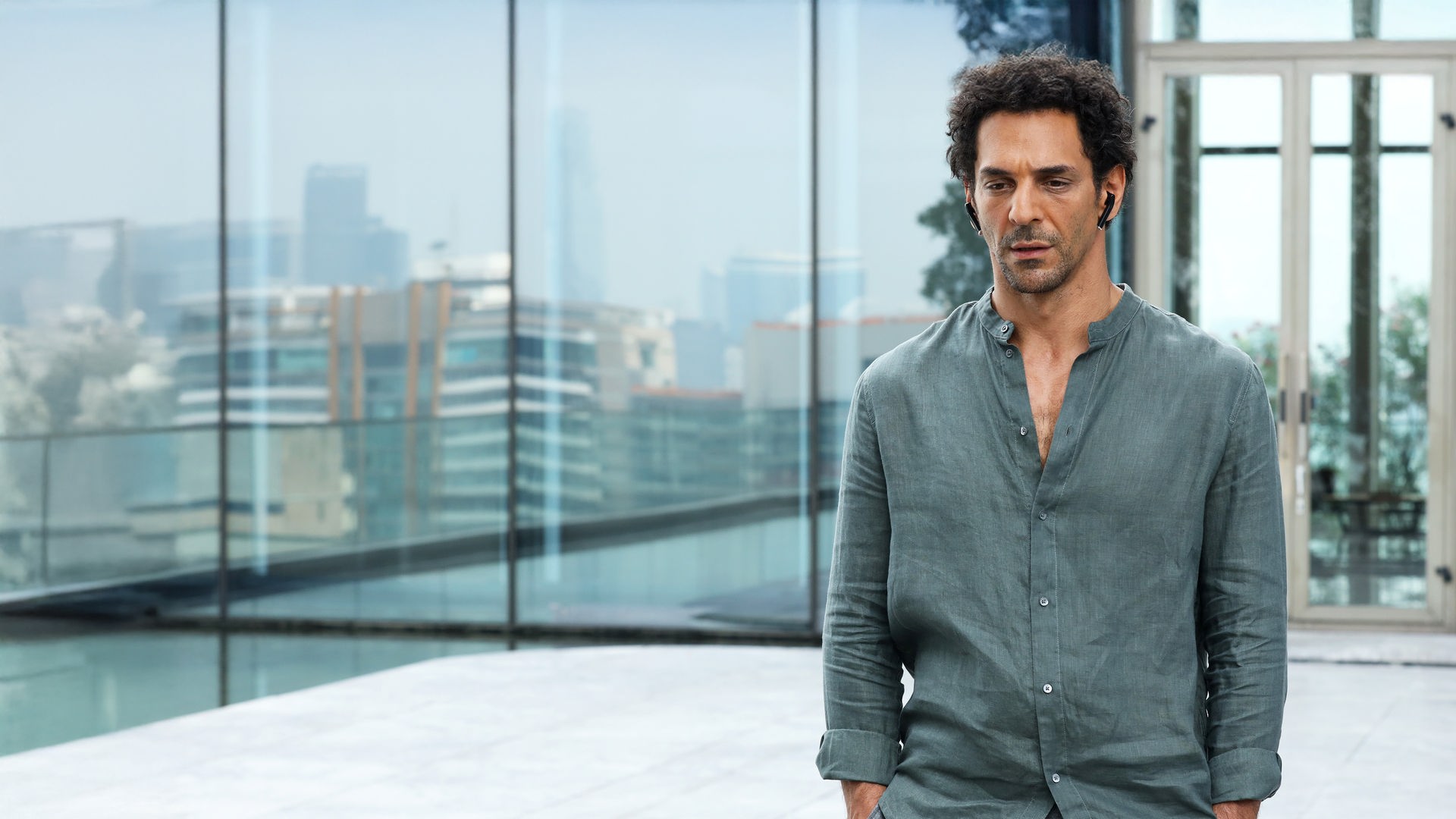 "Largo Winch - Der Preis des Geldes": Largo Winch (Tomer Sisley) blickt nachdenklich nach unten. Er trägt ein salbeigrünes Leinenhemd und steht vor einer modernen Glasfassade, in der sich eine unscharfe Skyline spiegelt.