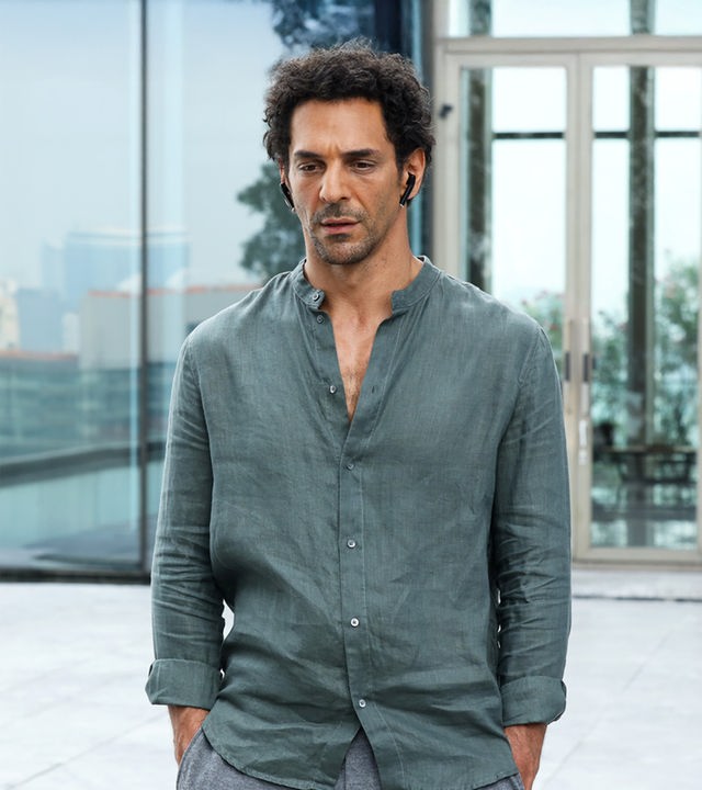 "Largo Winch - Der Preis des Geldes": Largo Winch (Tomer Sisley) blickt nachdenklich nach unten. Er trägt ein salbeigrünes Leinenhemd und steht vor einer modernen Glasfassade, in der sich eine unscharfe Skyline spiegelt.