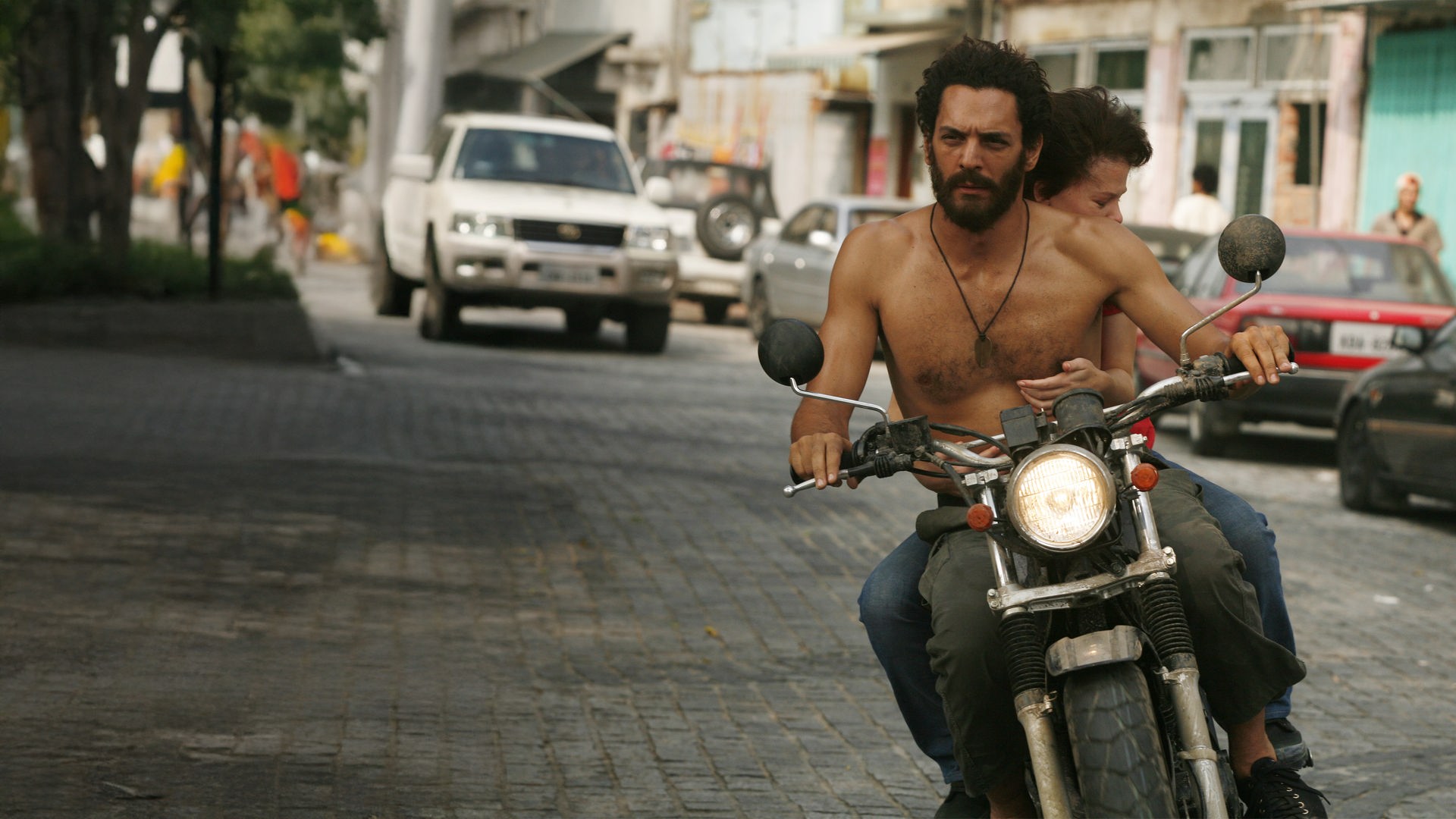 "Largo Winch - Tödliches Erbe": Largo (Tomer Sisley) fährt, Oberkörper frei, mit dem Motorrad durch die Stadt. Hinter ihm sitzt Lea (Mélanie Thierry) und hält sich an ihm fest.