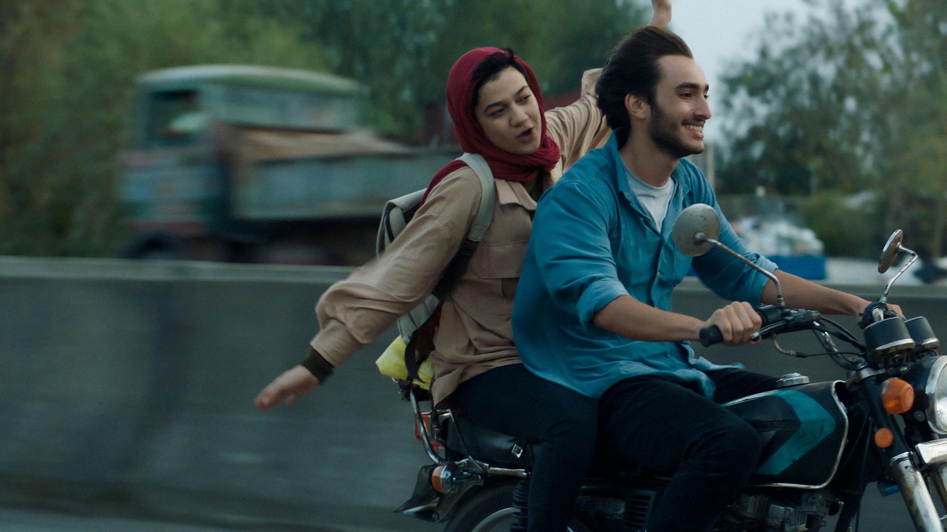 Narges (Sadaf Asgari) sitzt hinter Amir (Hamid Reza Abbasi) auf einem Motorrad und breitet die Arme au
