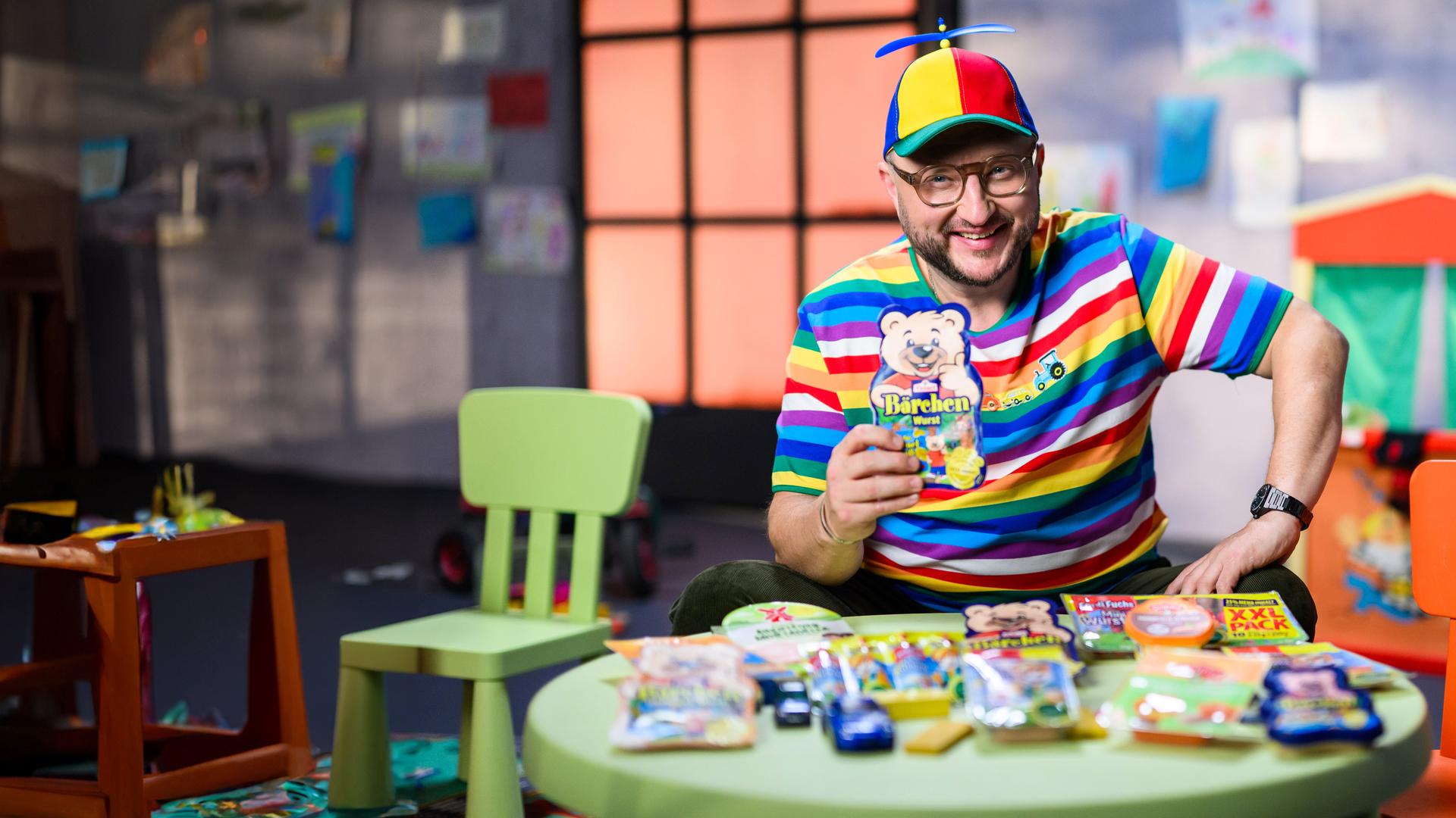 Sebastian Lege sitzt mit gestreiften Pullover und Propellermütze, mit einer Packung Bärchenwurst in der Hand auf einem Kinderstuhl.