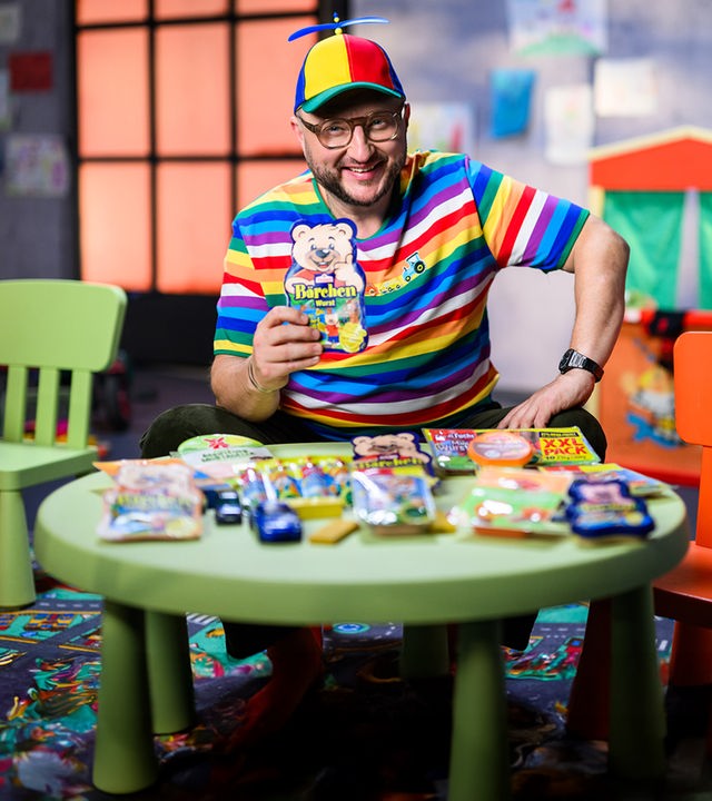 Sebastian Lege sitzt mit gestreiften Pullover und Propellermütze, mit einer Packung Bärchenwurst in der Hand auf einem Kinderstuhl.