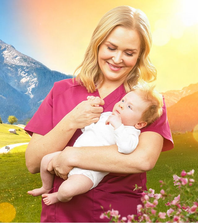 Hebamme Lena Lorenz (Judith Hoersch) steht vor der sonnigen Bergkulisse des Watzmann in den österreichischen Alpen. Sie hält ein Baby im Arm und schaut es liebevoll an.