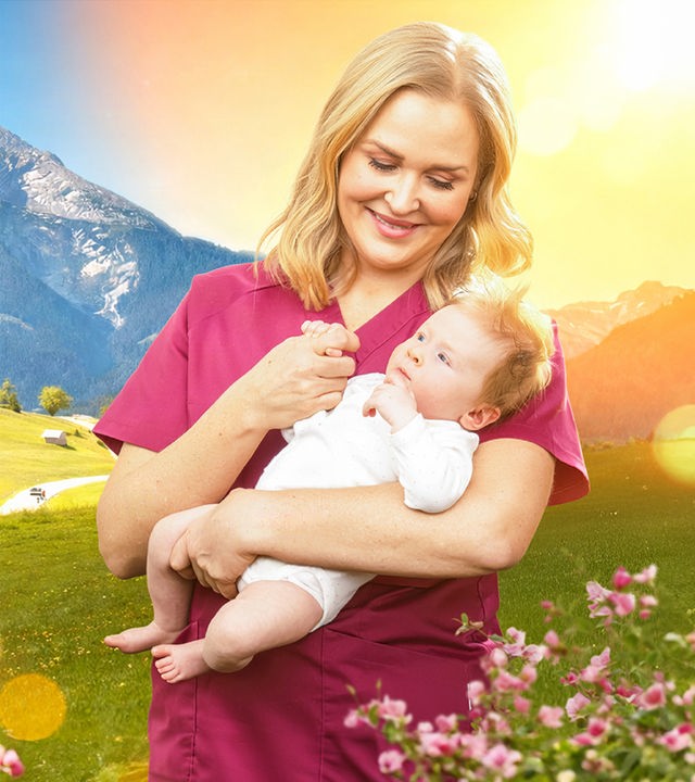 Hebamme Lena Lorenz (Judith Hoersch) steht vor der sonnigen Bergkulisse des Watzmann in den österreichischen Alpen. Sie hält ein Baby im Arm und schaut es liebevoll an.