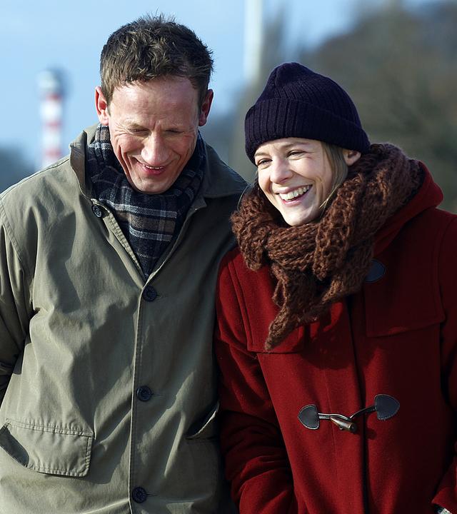 Leo Schreiber (links, Wotan Wilke Möhring) und Marie Johanson (rechts, Bernadette Heerwagen) spazieren gemeinsam am Elbstrand entlang. Sie laufen in wetterfester Kleidung nebeneinander und lachen glücklich. Links im Hintergrund erstreckt sich der Elbstrand.