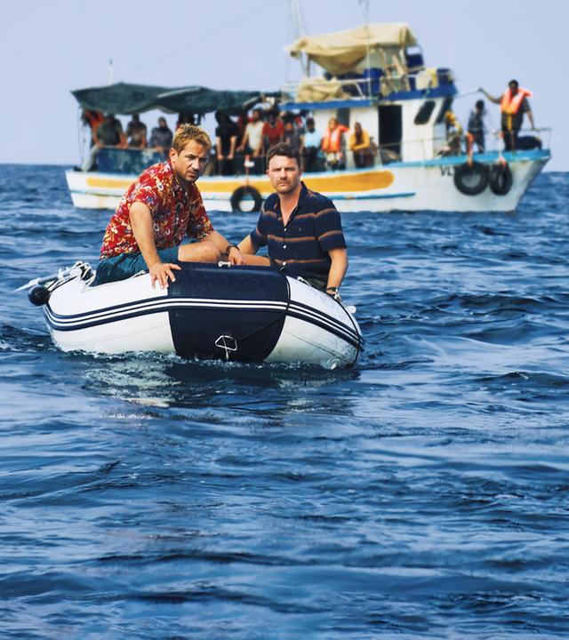 "LIBERAME - Nach dem Sturm - Abdrift": Mit einem Paddelboot fahren Daniel Schilling (Marc Benjamin) und Jan Garbe (Friedrich Mücke) auf dem Meer in Richtung Kamera. Im Hintergrund schwimmt ein Fischerboot.