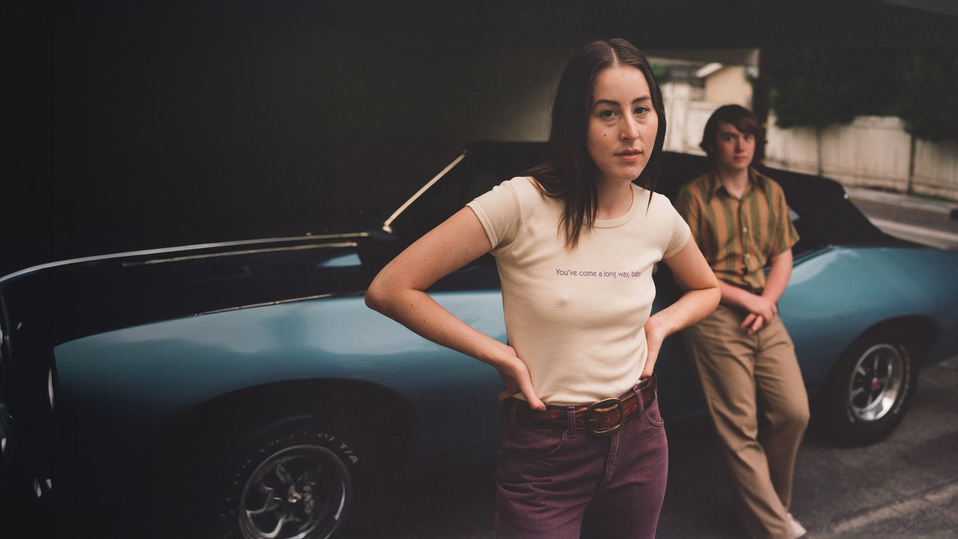 Das Filmplakat zu „Licorice Pizza“ zeigt die Hauptdarsteller Alana Haim und Cooper Hoffman im 70er-Jahre-Look, wie sie lässig vor einem blauen Oldtimer-Cabriolet posieren.