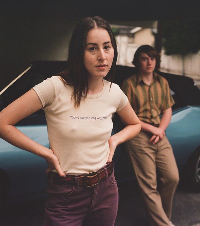 Das Filmplakat zu „Licorice Pizza“ zeigt die Hauptdarsteller Alana Haim und Cooper Hoffman im 70er-Jahre-Look, wie sie lässig vor einem blauen Oldtimer-Cabriolet posieren.