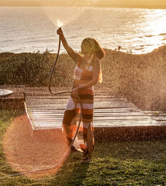 Clara (Penélope Cruz) steht mit einem über ihren Kopf erhobenen Gartenschlauch in einem Garten vor einer sommerlichen Seekulisse und spritzt Wasser in die Luft.