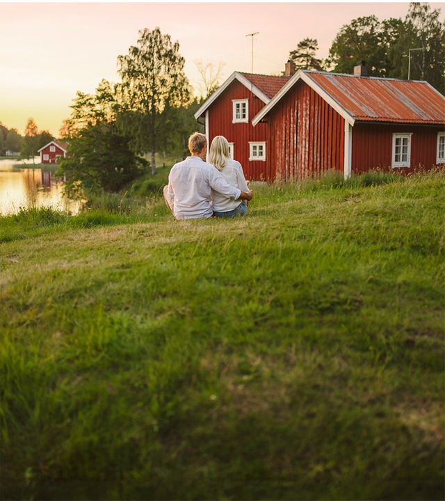 Ein verliebtes Paar sitzt im Gras am See und blickt auf ein typisch rotes Schwedenhäuschen