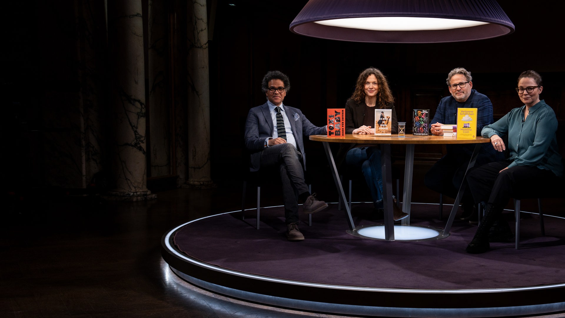 Ijoma Mangold, Sandra Richter, Wolfram Eilenberger und Gastgeberin Thea Dorn sitzen in einem dunklen Raum an einem Tisch und blicken in die Kamera. Sie werden von oben von einer Lampe beleuchtet.
