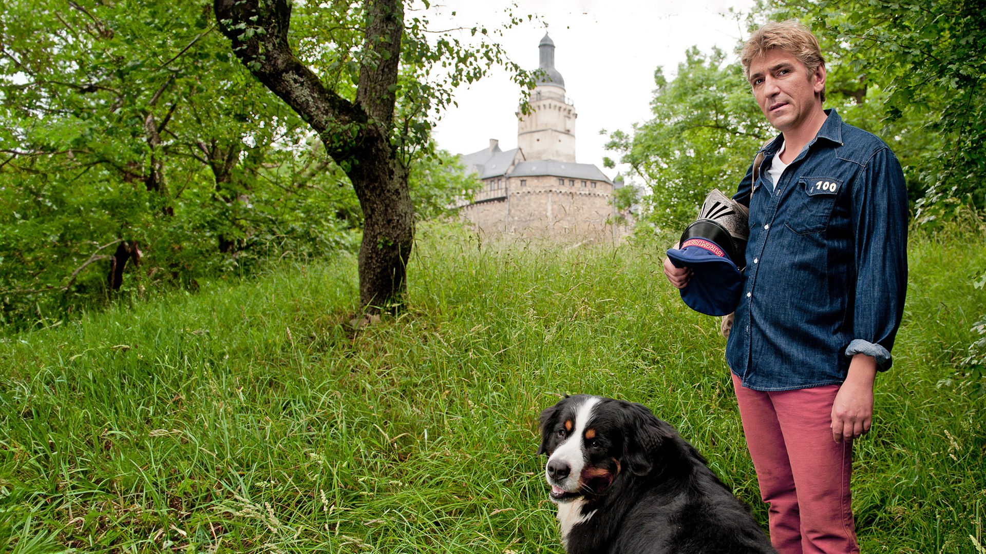 Fritz, mit einem Ritterhelm in der Hand, steht mit Keks auf einer Wiese - im Hintergrund ist eine Burg zu sehen