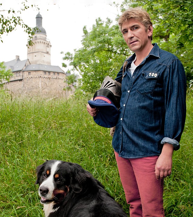 Fritz, mit einem Ritterhelm in der Hand, steht mit Keks auf einer Wiese - im Hintergrund ist eine Burg zu sehen