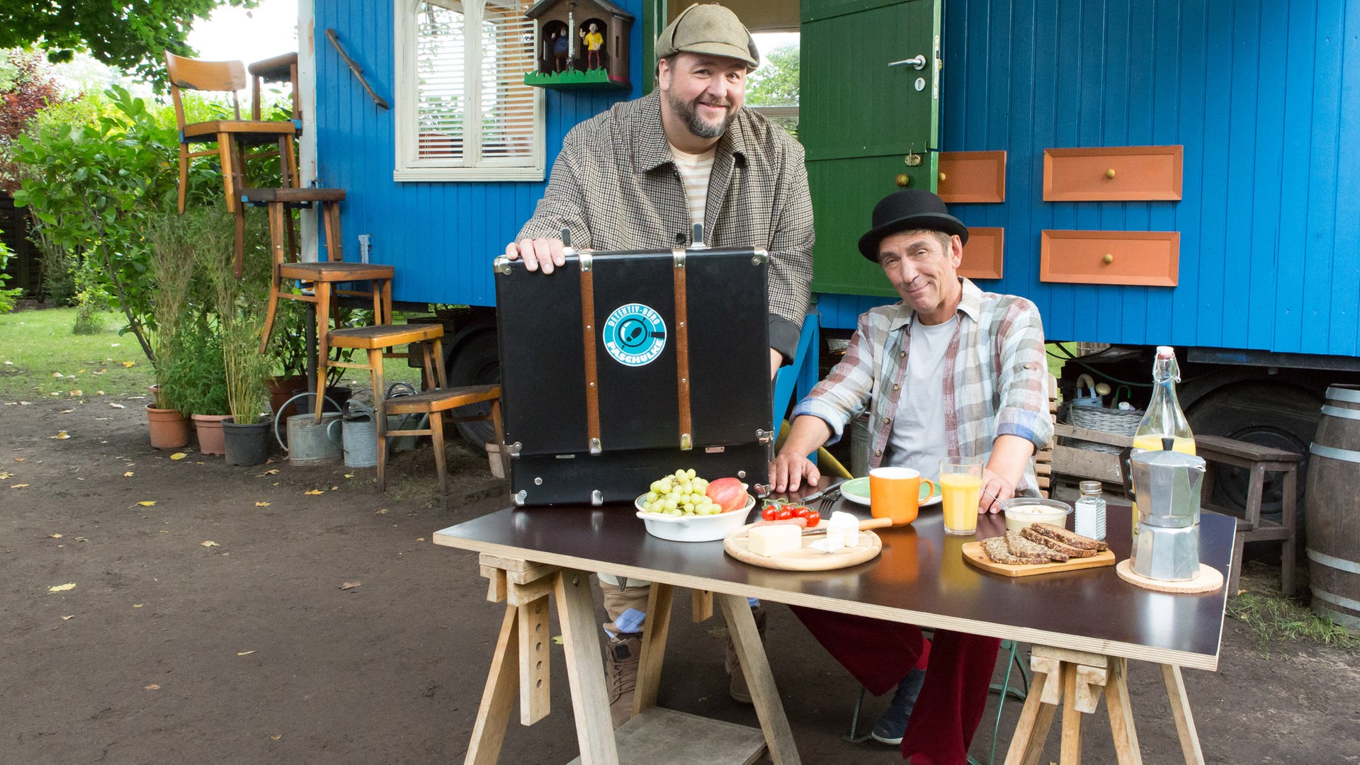 Fritz und Herr Paschulke sitzen bzw. stehen als Detektive verkleidet am Frühstückstisch vor dem Bauwagen