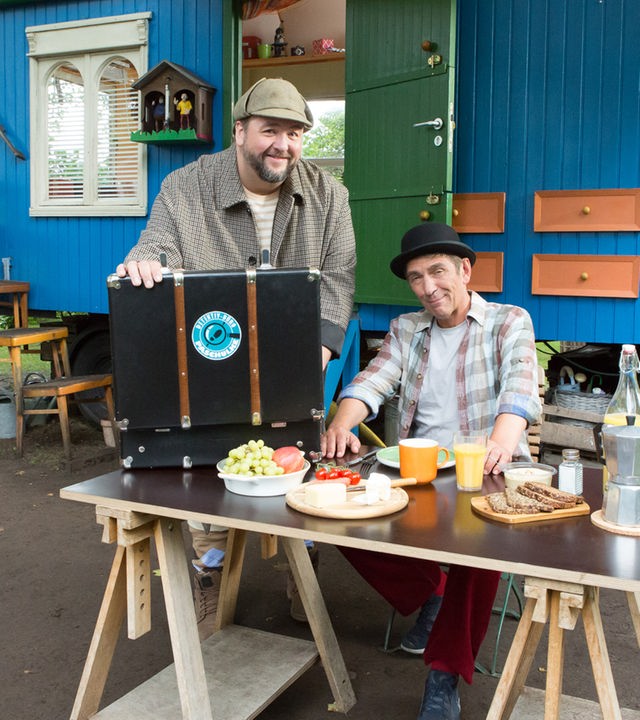 Fritz und Herr Paschulke sitzen bzw. stehen als Detektive verkleidet am Frühstückstisch vor dem Bauwagen