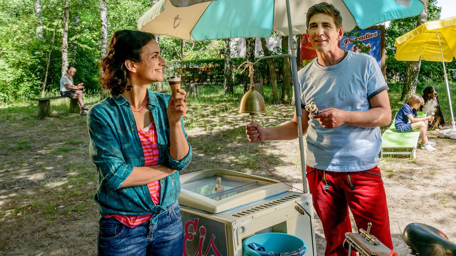 Fritz Fuchs hält ein Eis in der Hand und leutet eine Glocke von einem Eisverkaufsstand, neben ihm steht Yasemin