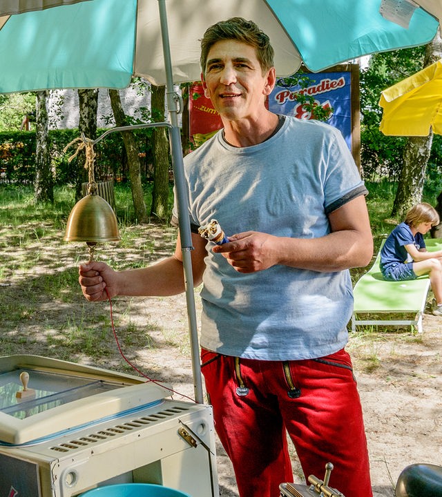 Fritz Fuchs hält ein Eis in der Hand und leutet eine Glocke von einem Eisverkaufsstand, neben ihm steht Yasemin