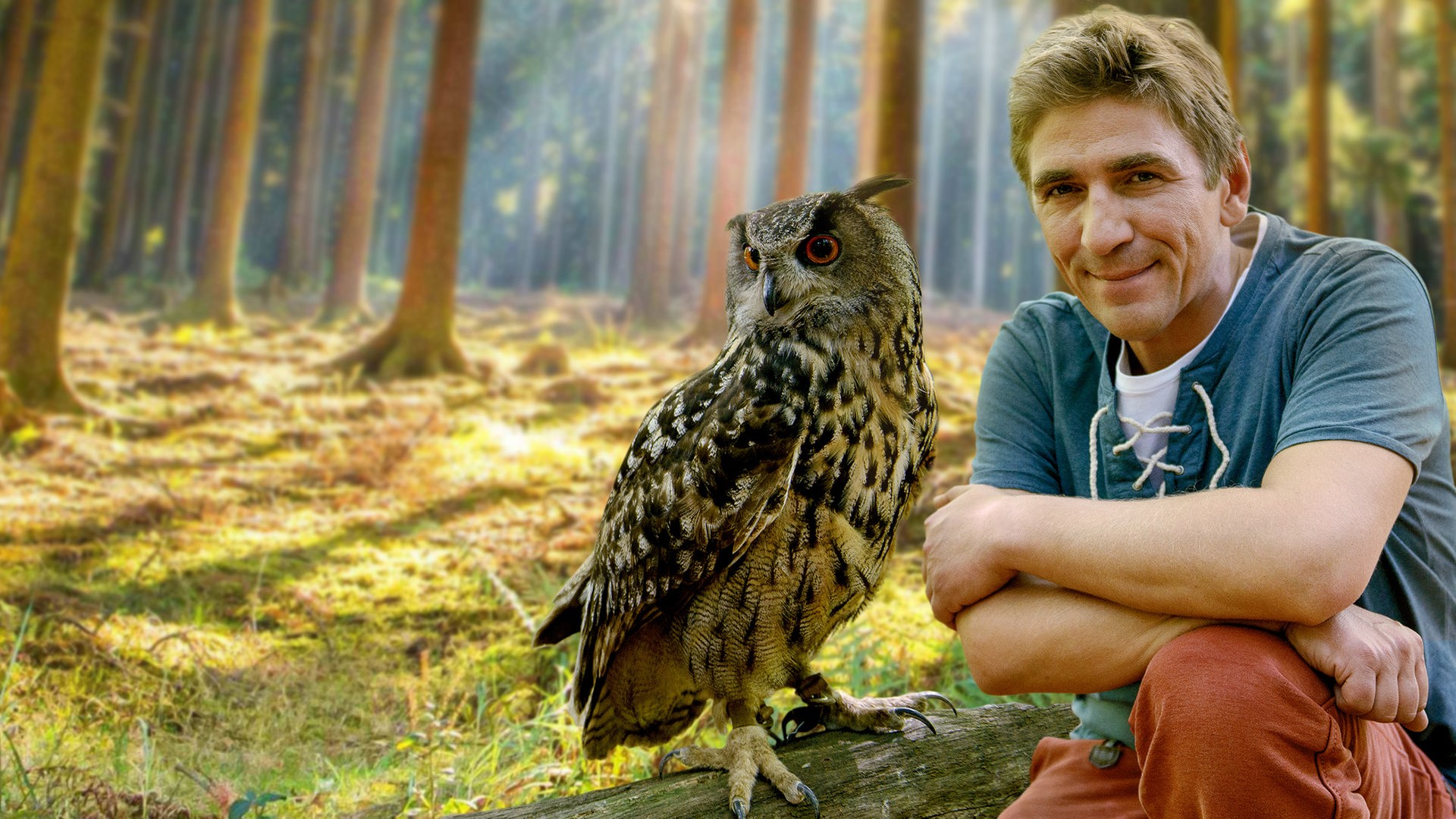 Fritz Fuchs hockt im Wald neben einer Eule, die auf einem am Boden liegenden Baumstamm sitzt