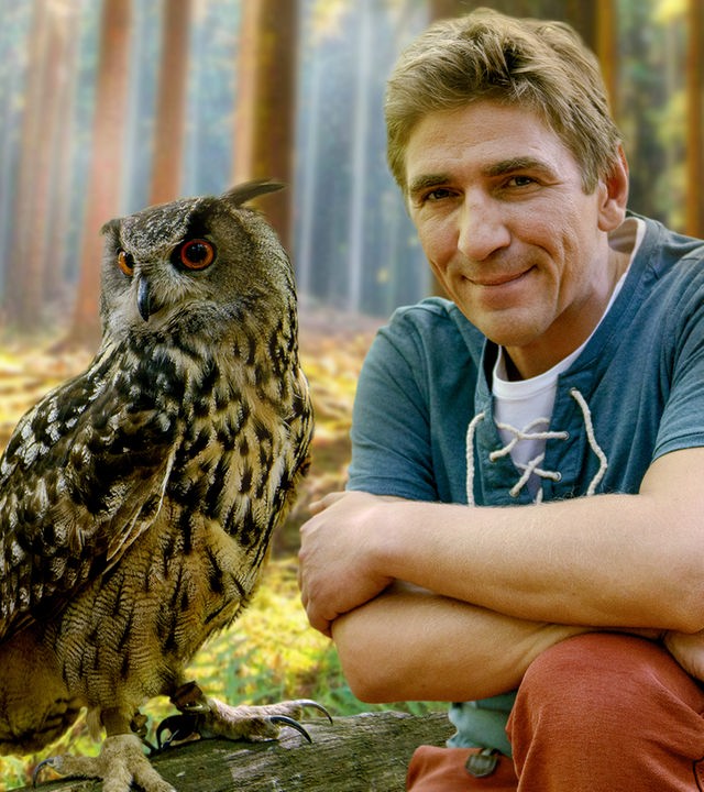 Fritz Fuchs hockt im Wald neben einer Eule, die auf einem am Boden liegenden Baumstamm sitzt