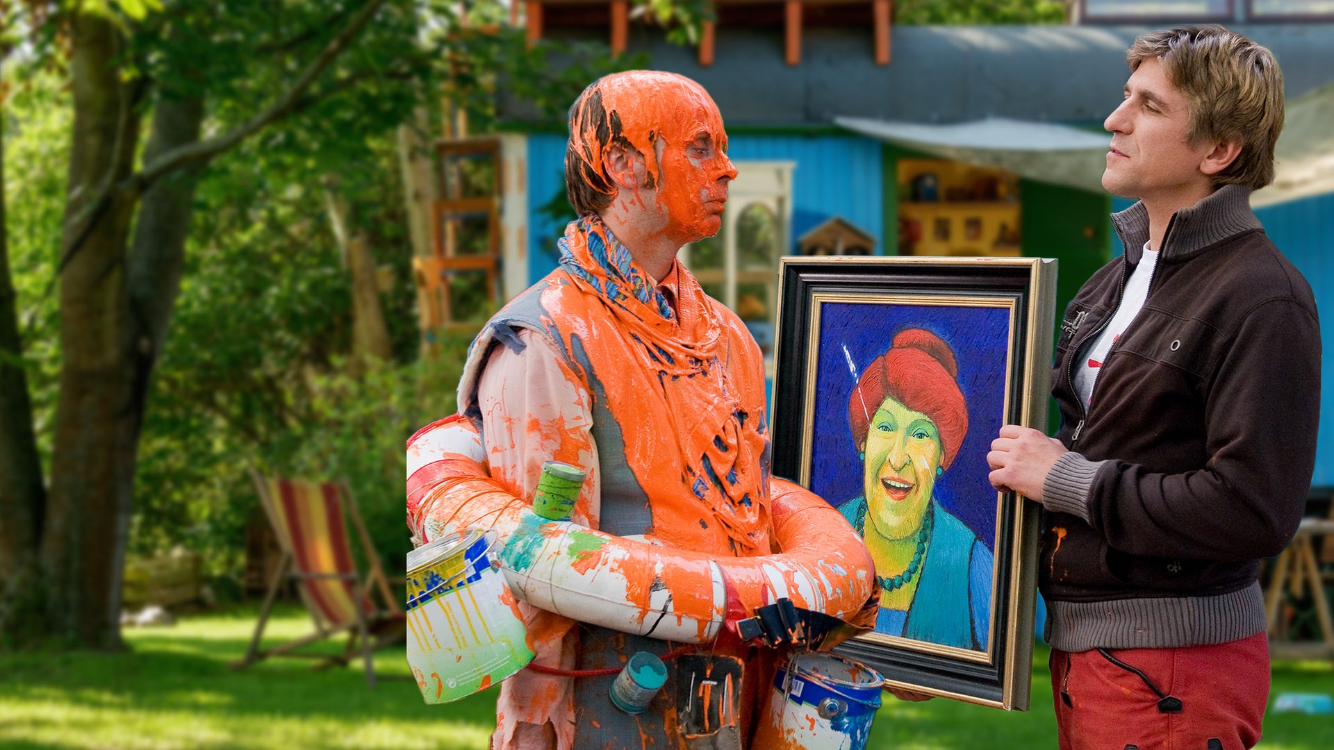 Fritz steht vor dem Bauwagen und hält ein Gemälde hoch - neben ihm steht ein Mann, der voller orangener Farbe ist