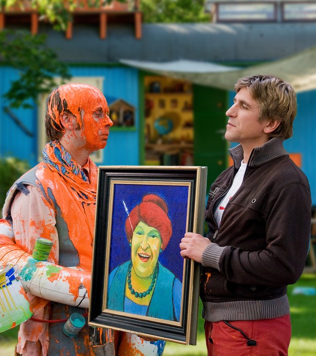 Fritz steht vor dem Bauwagen und hält ein Gemälde hoch - neben ihm steht ein Mann, der voller orangener Farbe ist