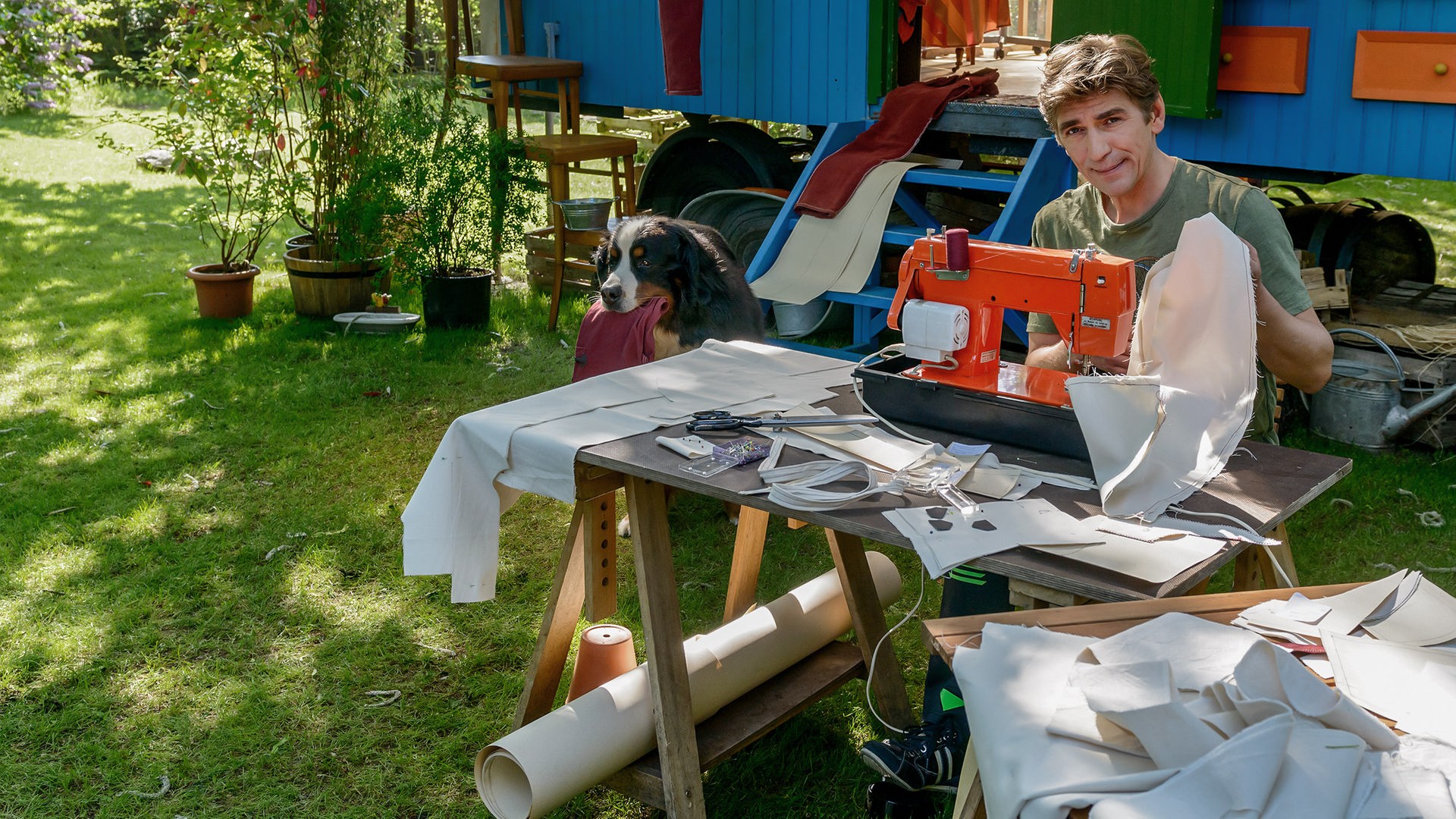 Fritz sitzt vor dem Bauwagen und arbeitet an einer Nähmaschine