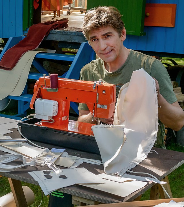 Fritz sitzt vor dem Bauwagen und arbeitet an einer Nähmaschine