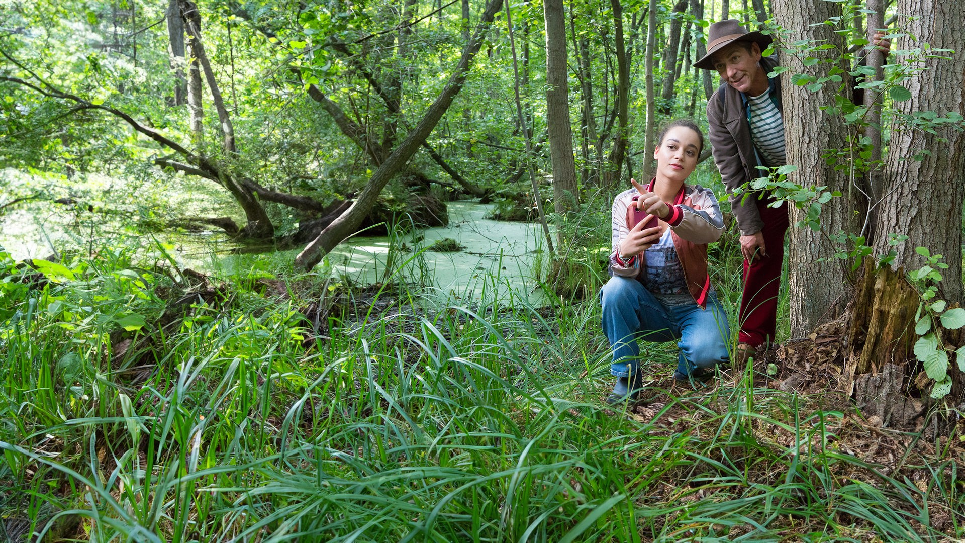 Leyla und Fritz befinden sich im Wald und beobachten etwas - im Hintergrund eine Moorlandschaft