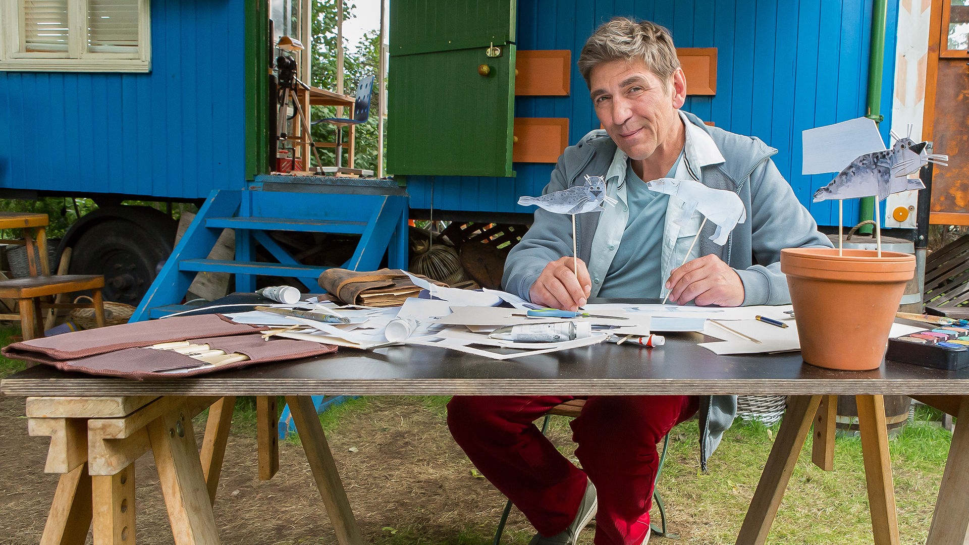 Fritz sitzt an einem Tisch vor dem Bauwagen und hält eine Papierrobbe hoch