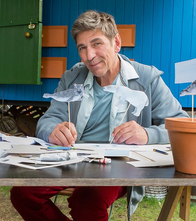 Fritz sitzt an einem Tisch vor dem Bauwagen und hält eine Papierrobbe hoch