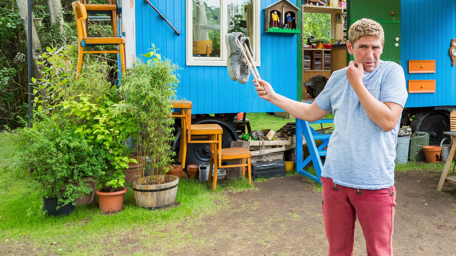 Fritz steht vor dem Bauwagen und hält mit Stöcken aufgespießte stinkende Schuhe hoch