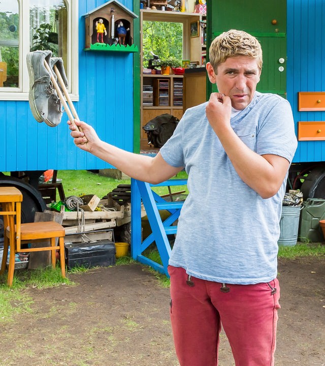 Fritz steht vor dem Bauwagen und hält mit Stöcken aufgespießte stinkende Schuhe hoch