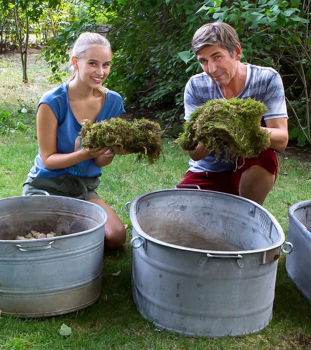 Fritz und Kim hocken vor mehreren großen Bottichen und halten jeweils einen Moosballen hoch