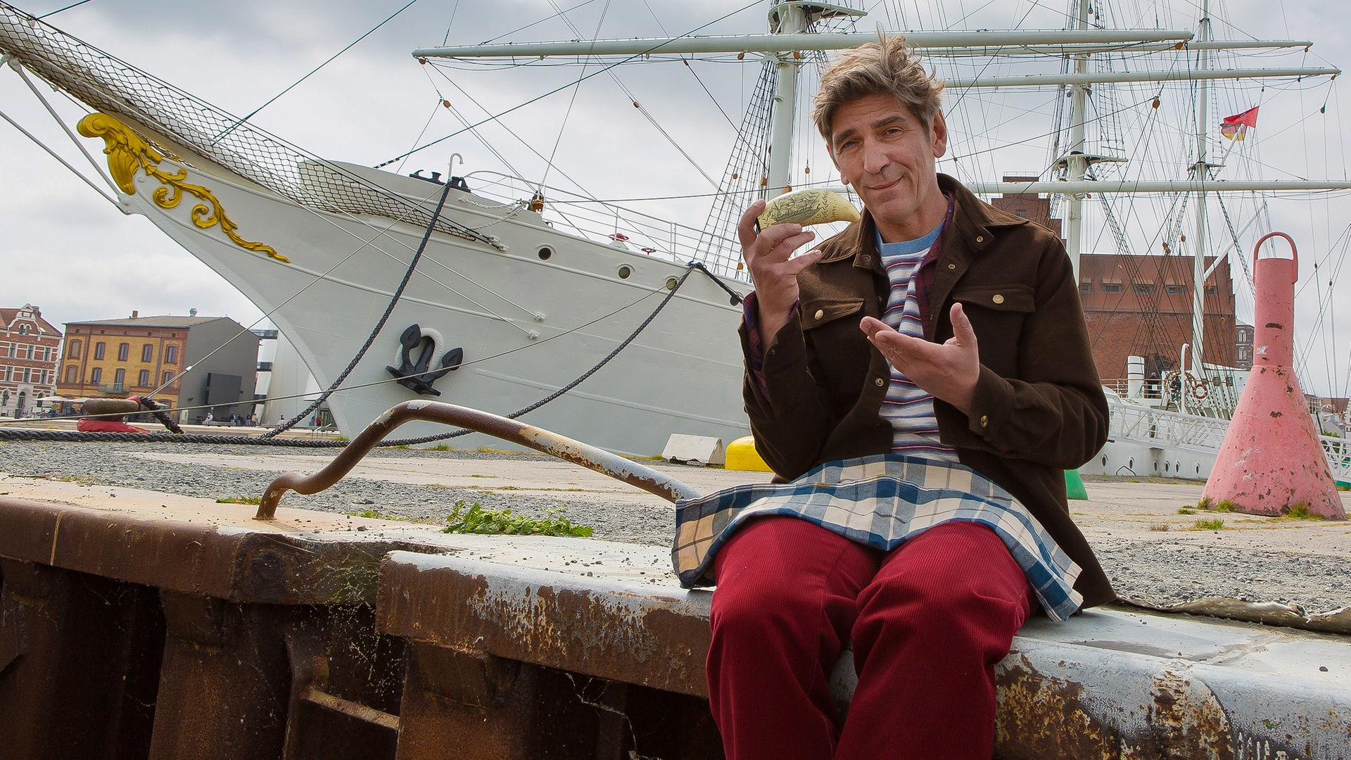 Fritz sitzt auf einer Kaimauer und hat einen Walzahn in der Hand - im Hintergrund liegt ein Segelschiff im Hafen
