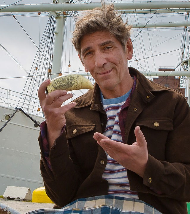 Fritz sitzt auf einer Kaimauer und hat einen Walzahn in der Hand - im Hintergrund liegt ein Segelschiff im Hafen
