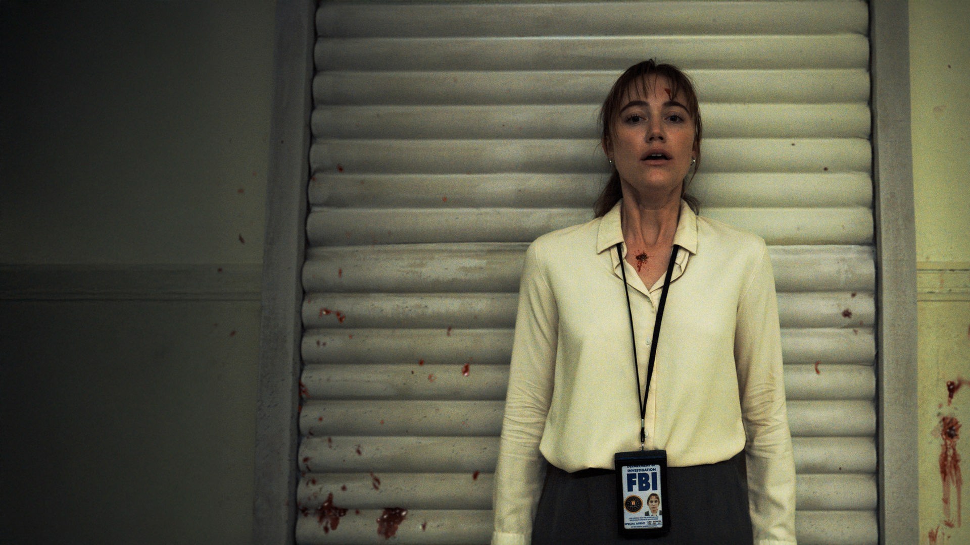"Longlegs": Lee Harker (Maika Monroe) lehnt mit dem Rücken an einer Tür und schaut schockiert nach vorne. Blutspuren zeichnen sich auf ihrer Stirn und an ihrem Hals ab, weitere Spritzer befinden sich an der Wand neben ihr.