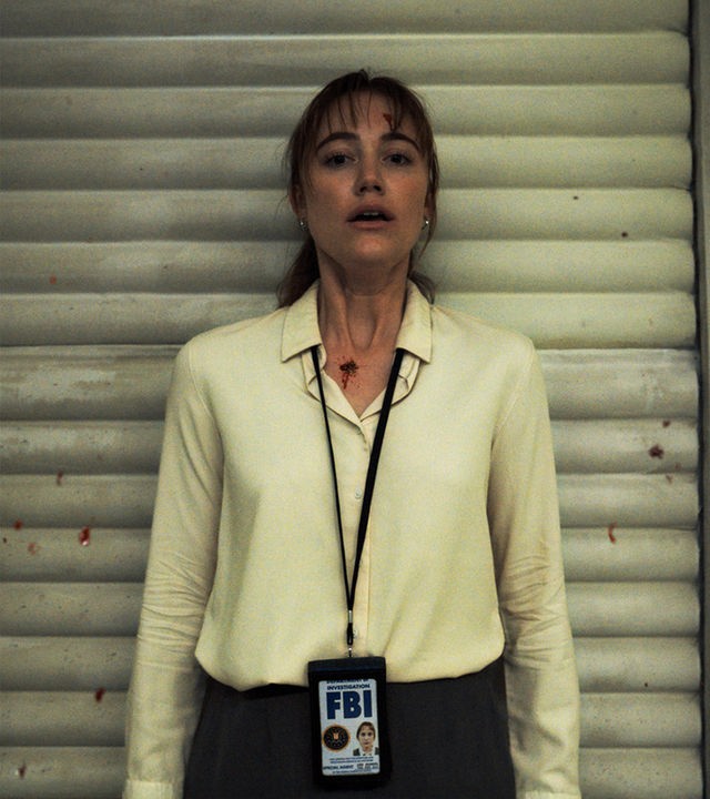 "Longlegs": Lee Harker (Maika Monroe) lehnt mit dem Rücken an einer Tür und schaut schockiert nach vorne. Blutspuren zeichnen sich auf ihrer Stirn und an ihrem Hals ab, weitere Spritzer befinden sich an der Wand neben ihr.